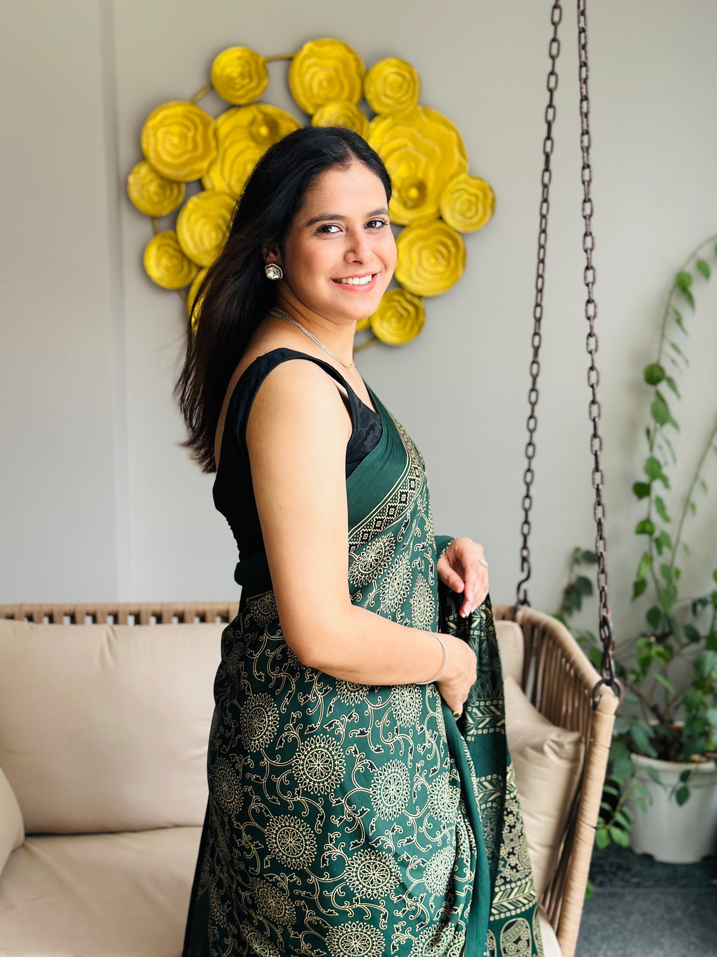 Woman in a green saree standing in a room with decorative wall art and plants.