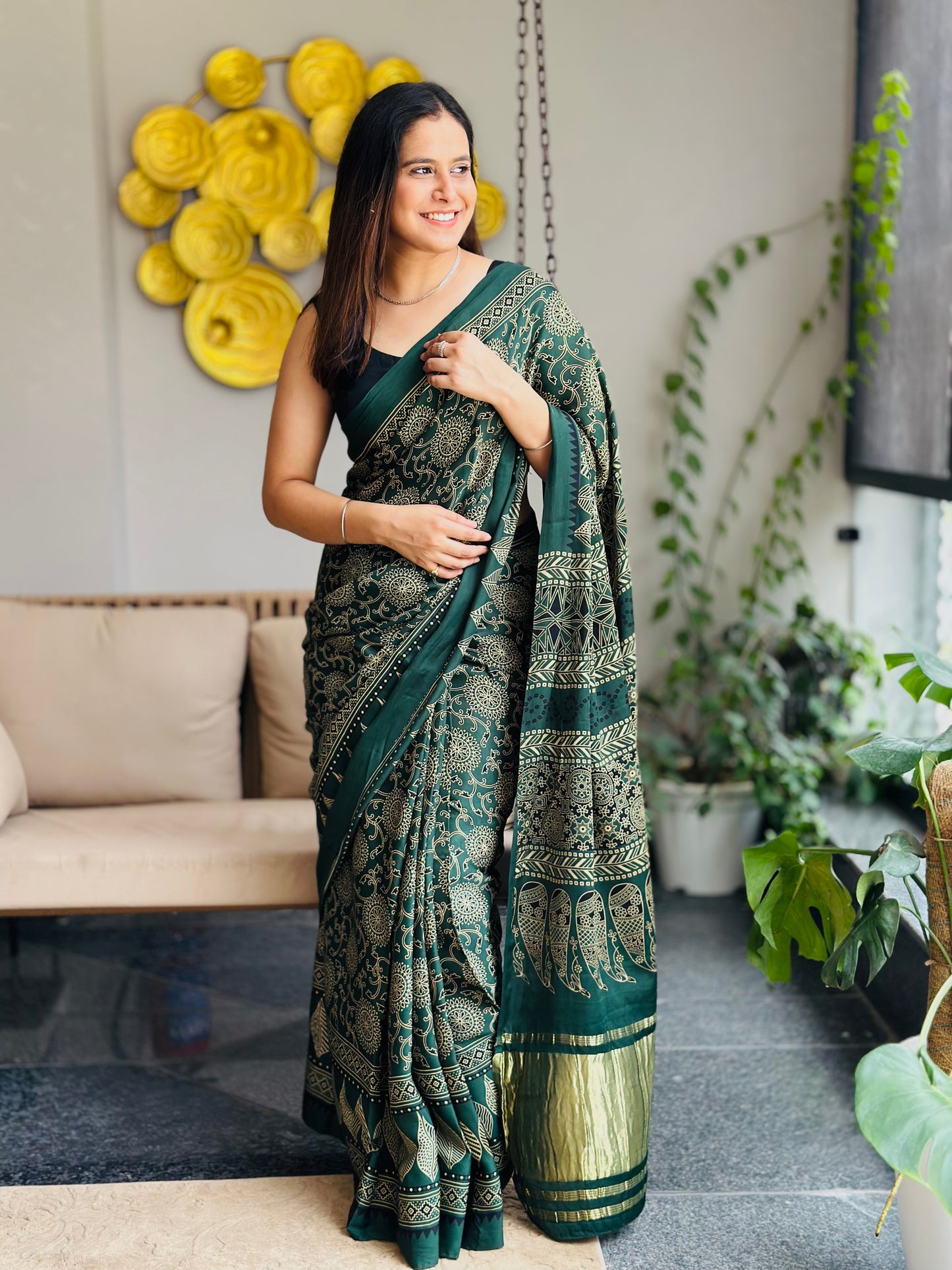 Woman in a green saree standing in a living room with plants and a beige sofa.