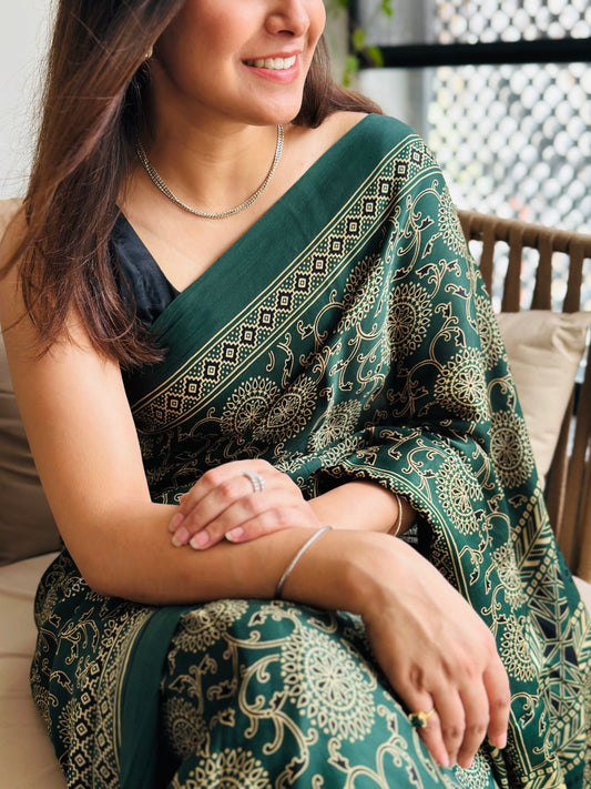 Woman wearing a green saree with intricate patterns, sitting outdoors.